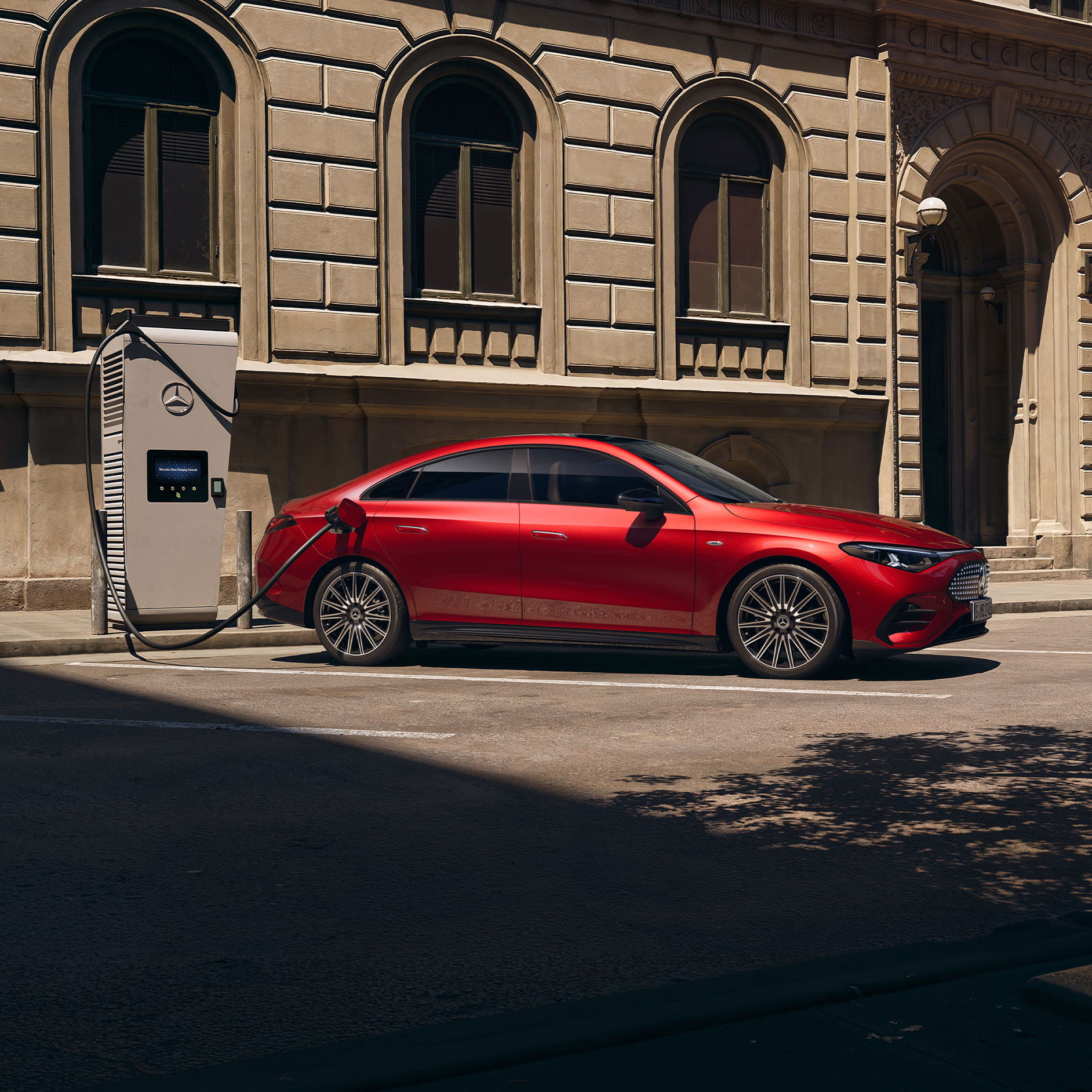 Röd Mercedes-Benz CLA Electric ansluten till en laddstation, parkerad vid trottoarkanten.