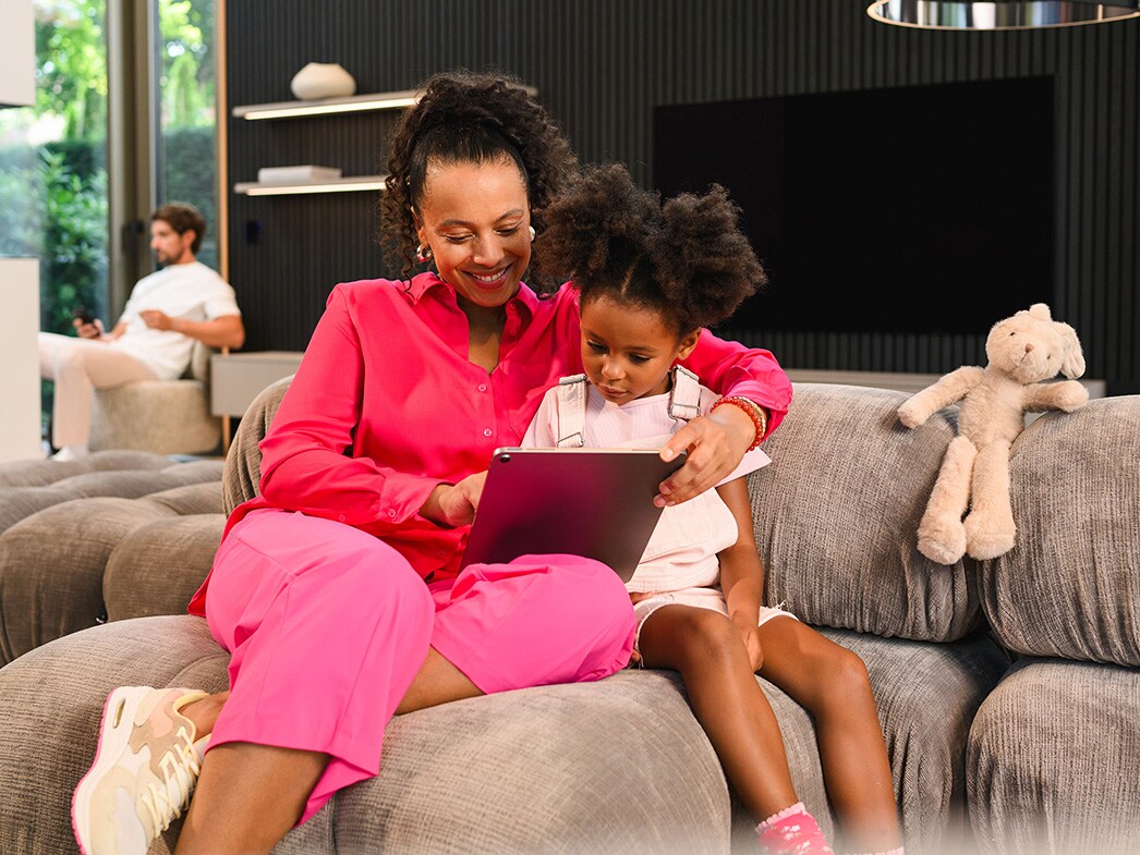 Fördelarna med Mercedes-Benz Finance | Mercedes-Benz Transportbilar En kvinna och ett barn sitter i en soffa och använder en surfplatta, medan en man sitter i bakgrunden.