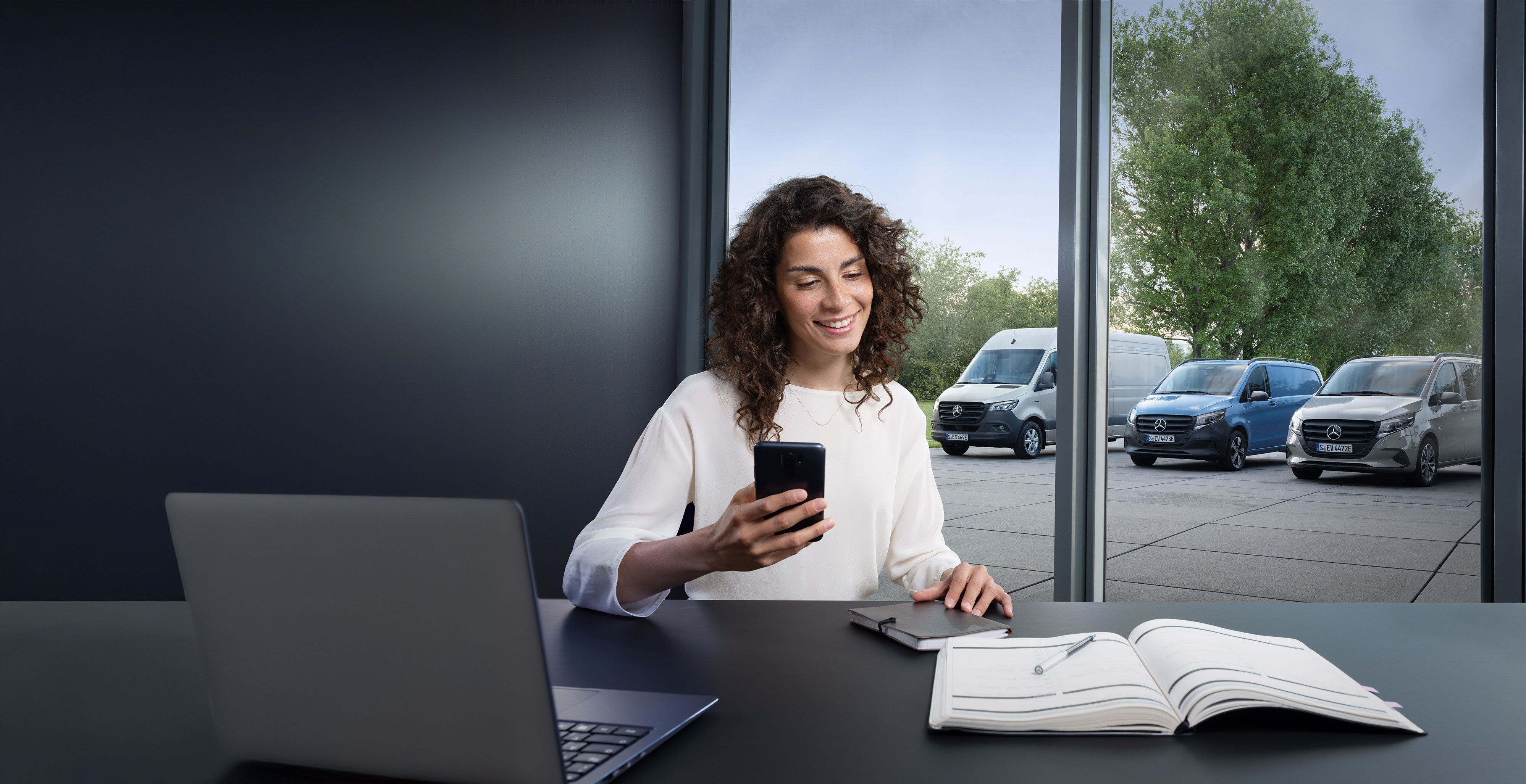 Mercedes-Benz | Vans Care Kvinna med smartphone vid ett skrivbord, flera Mercedes-Benz transportbilar i bakgrunden – experthjälp inom företagsservice.